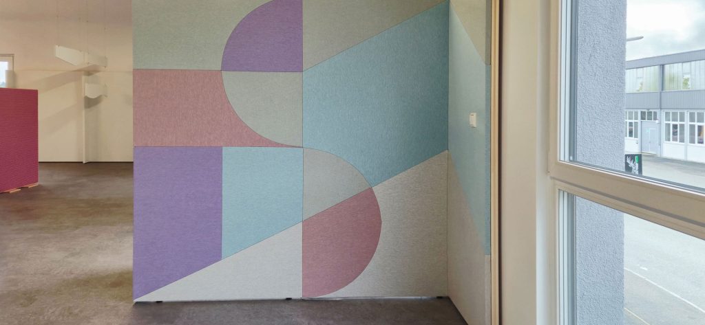 A modern interior wall featuring a geometric mural with pastel shapes in blue, purple, pink, and gray. The design includes circles, triangles, and rectangles. Large windows are visible to the right, letting in natural light.