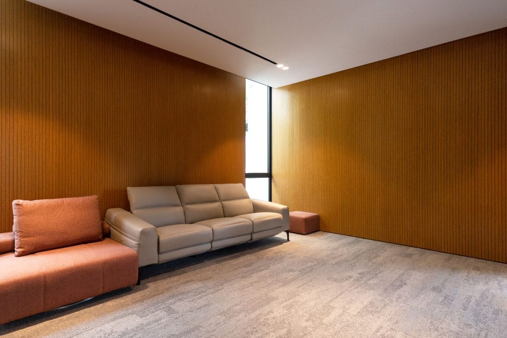 A modern room with light gray carpet, wood-paneled walls, a beige leather sofa, two brown upholstered chairs, and a matching ottoman near a tall, narrow window letting in natural light.