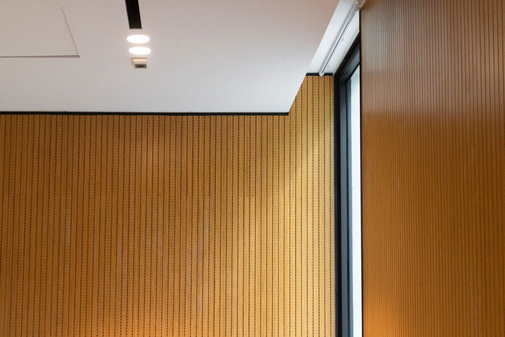 A modern interior with light brown, vertically-striped wooden wall panels, a large window on the right, and a white ceiling with recessed lights.