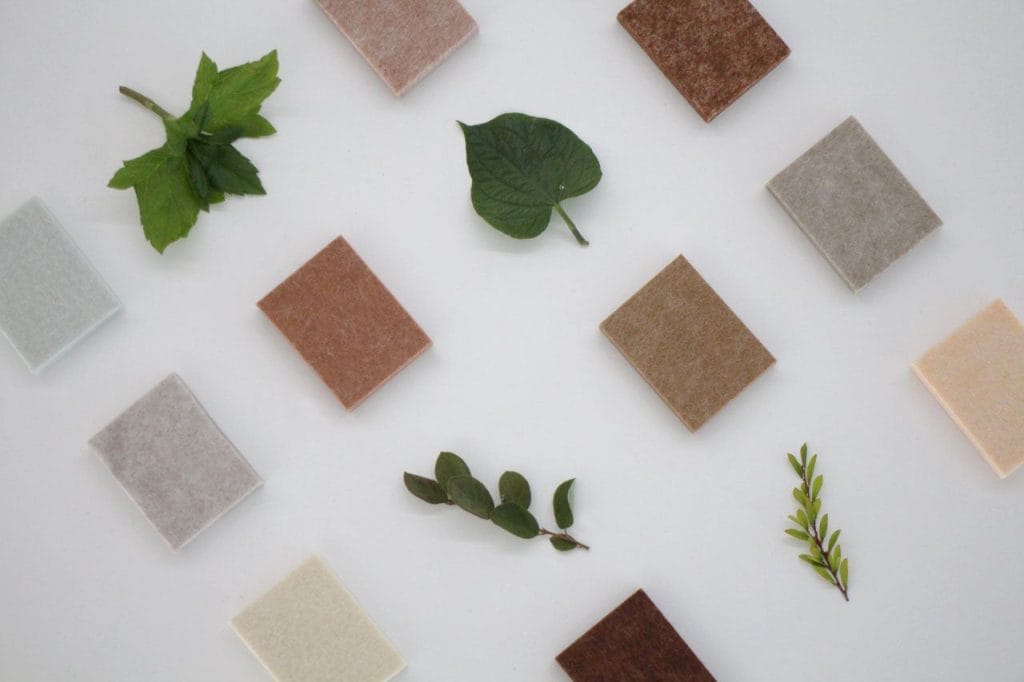 A flat lay of square fabric swatches in neutral tones arranged around assorted green leaves on a white background.