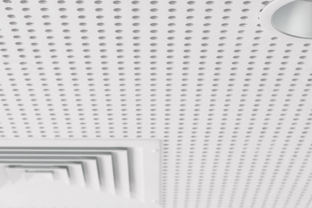 White perforated ceiling with circular holes, a recessed light fixture in the upper right corner, and a square air vent with layered slats in the lower left corner. The overall appearance is clean and minimalistic.