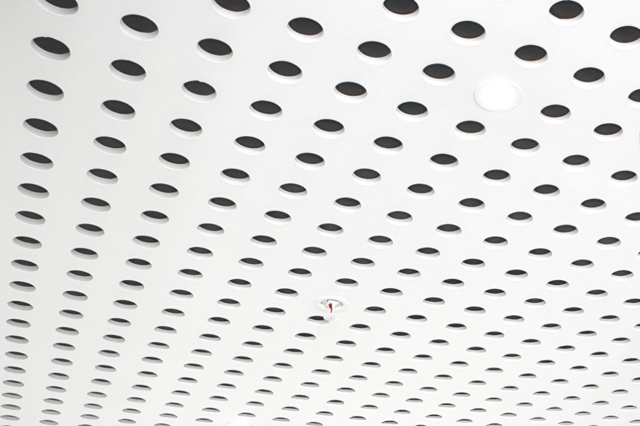 A white ceiling with a grid of evenly spaced circular holes and a small red fire sprinkler in the center.