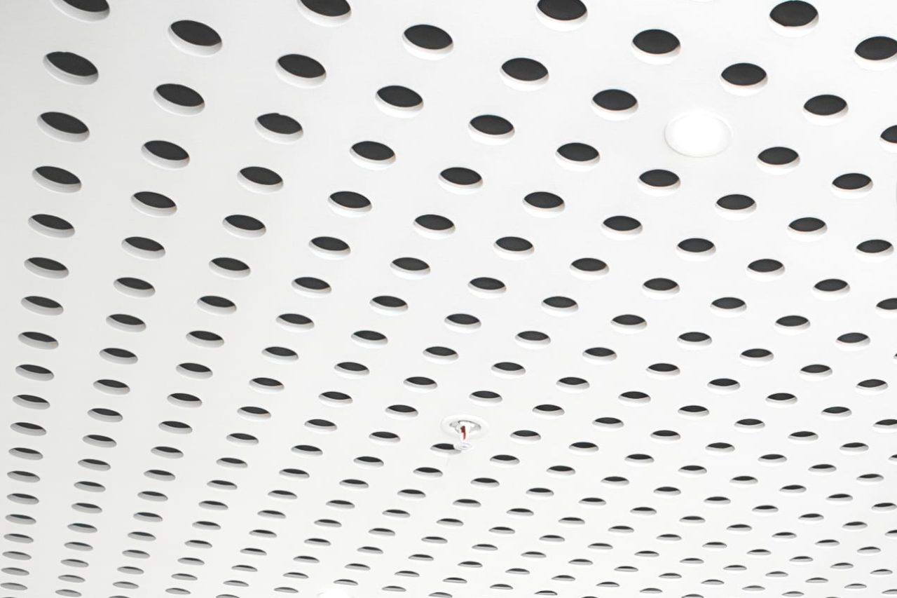 A white ceiling with a grid of evenly spaced circular holes and a small red fire sprinkler in the center.