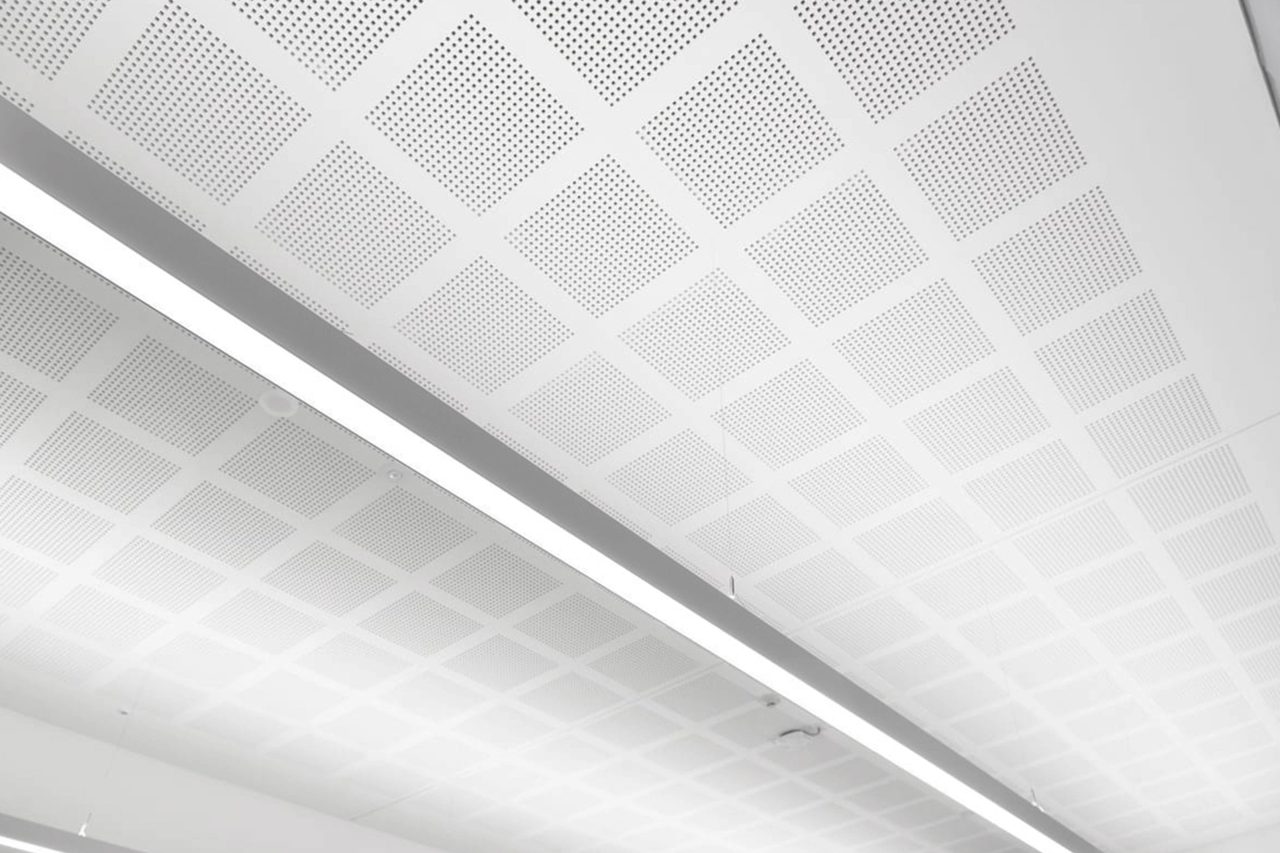 A white ceiling with a geometric pattern of perforated square panels and two long, narrow fluorescent light fixtures running parallel to each other.