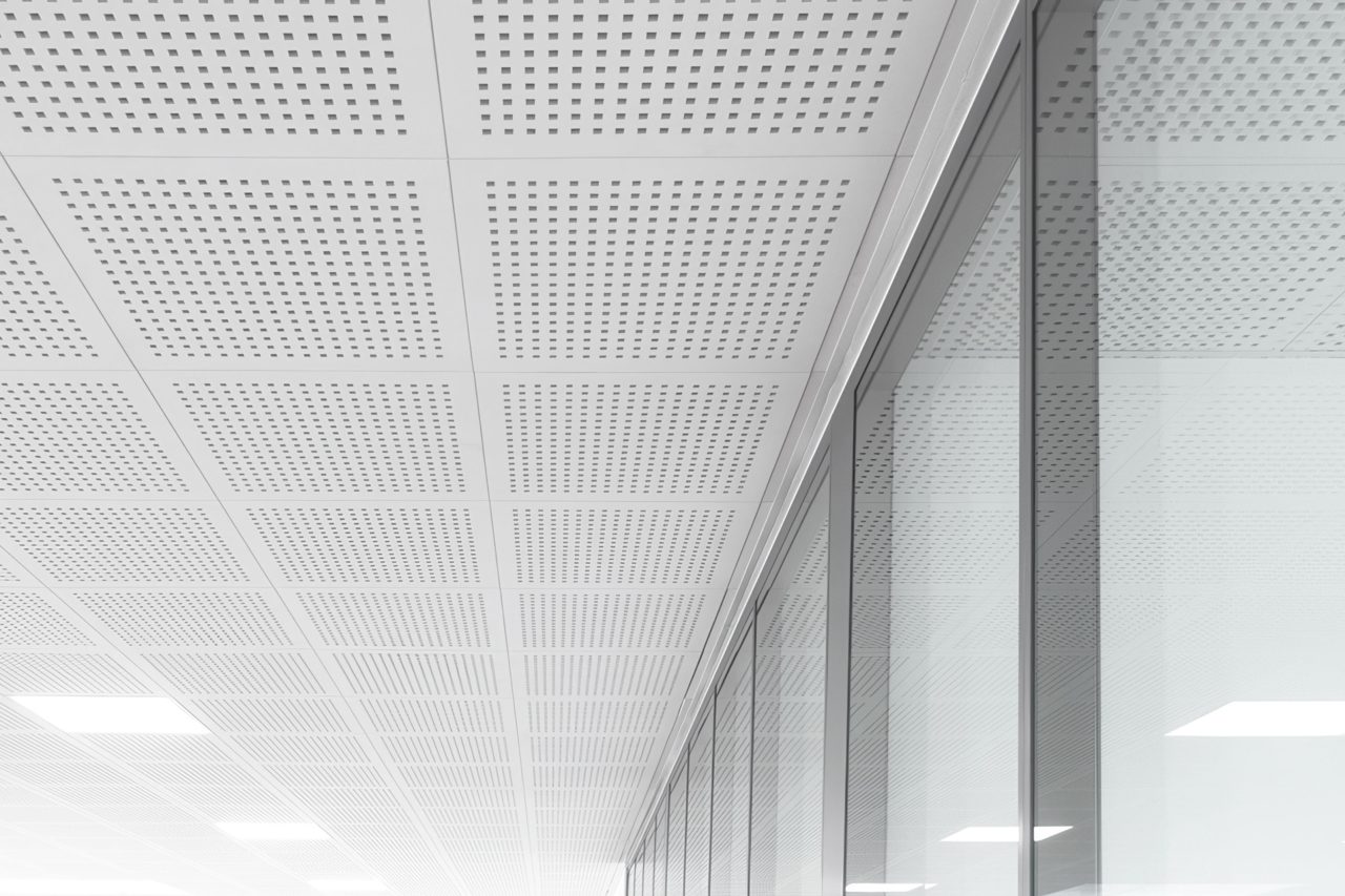 A modern, minimalist ceiling with square perforated panels and bright lighting, reflected in a large glass wall on the right side of the image. The scene is clean, sleek, and mostly white.