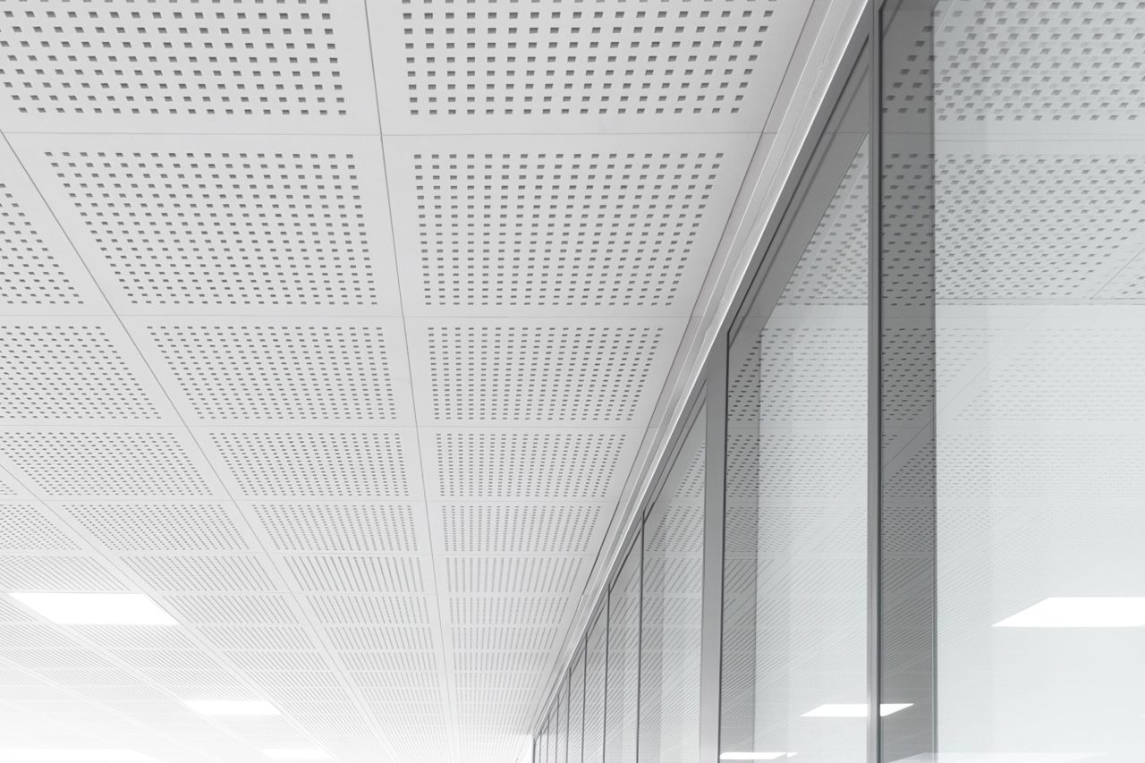 A modern, minimalist ceiling with square perforated panels and bright lighting, reflected in a large glass wall on the right side of the image. The scene is clean, sleek, and mostly white.