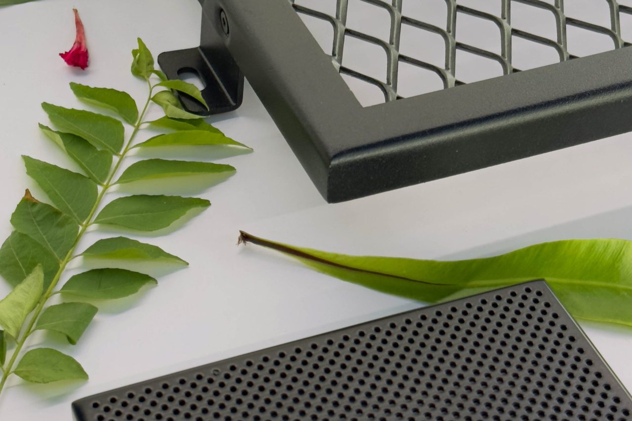 Several green leaves are arranged on a white surface next to a metal grid and a black perforated rectangular object, possibly electronic or automotive components.