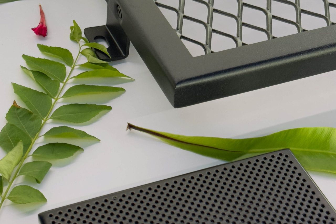 Several green leaves are arranged on a white surface next to a metal grid and a black perforated rectangular object, possibly electronic or automotive components.