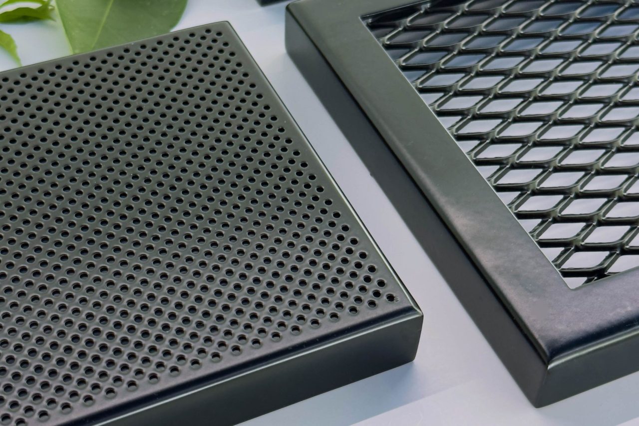 Three black metal air vent covers, one with round holes and another with a diamond mesh pattern, are placed on a surface with green leaves and small red flowers scattered around.