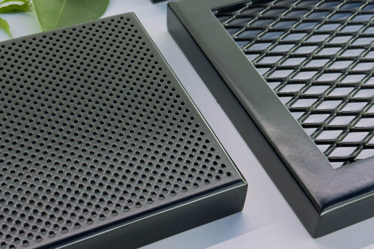 Three black metal air vent covers, one with round holes and another with a diamond mesh pattern, are placed on a surface with green leaves and small red flowers scattered around.