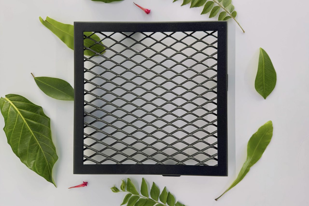 A black, square metal mesh panel is placed on a white surface, surrounded by various green leaves and a small red flower. The mesh has a diamond-shaped grid pattern.