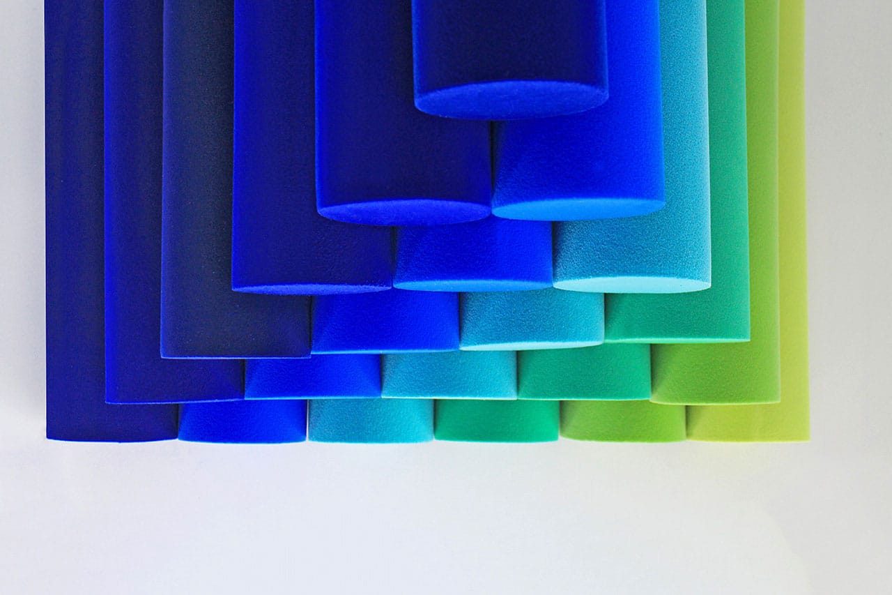 Cylindrical foam tubes in gradient shades of blue, teal, and green are arranged vertically and overlap each other against a white background.