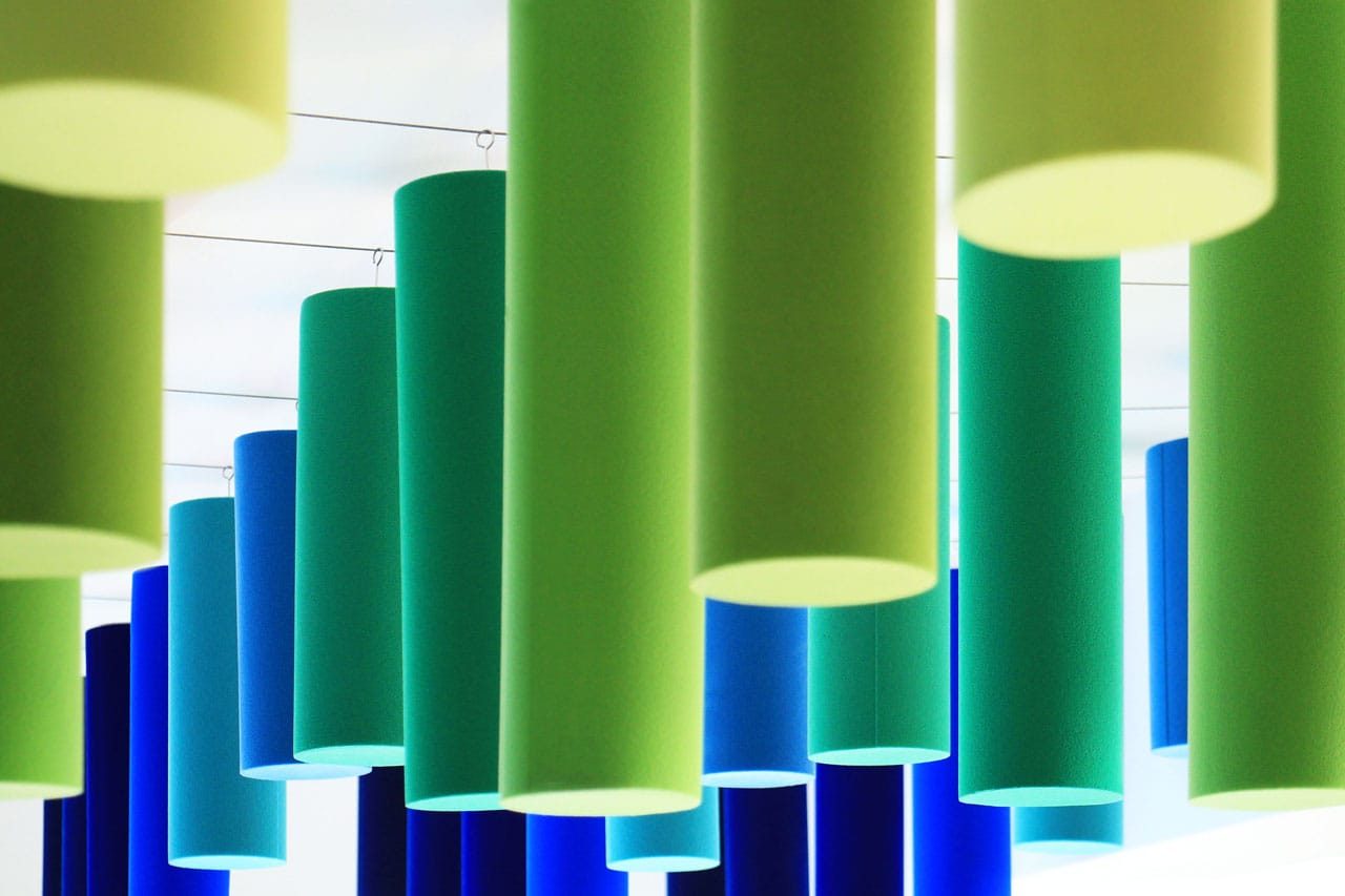 Cylindrical hanging lights in shades of green and blue are suspended from the ceiling in rows, creating a colorful and modern geometric pattern.