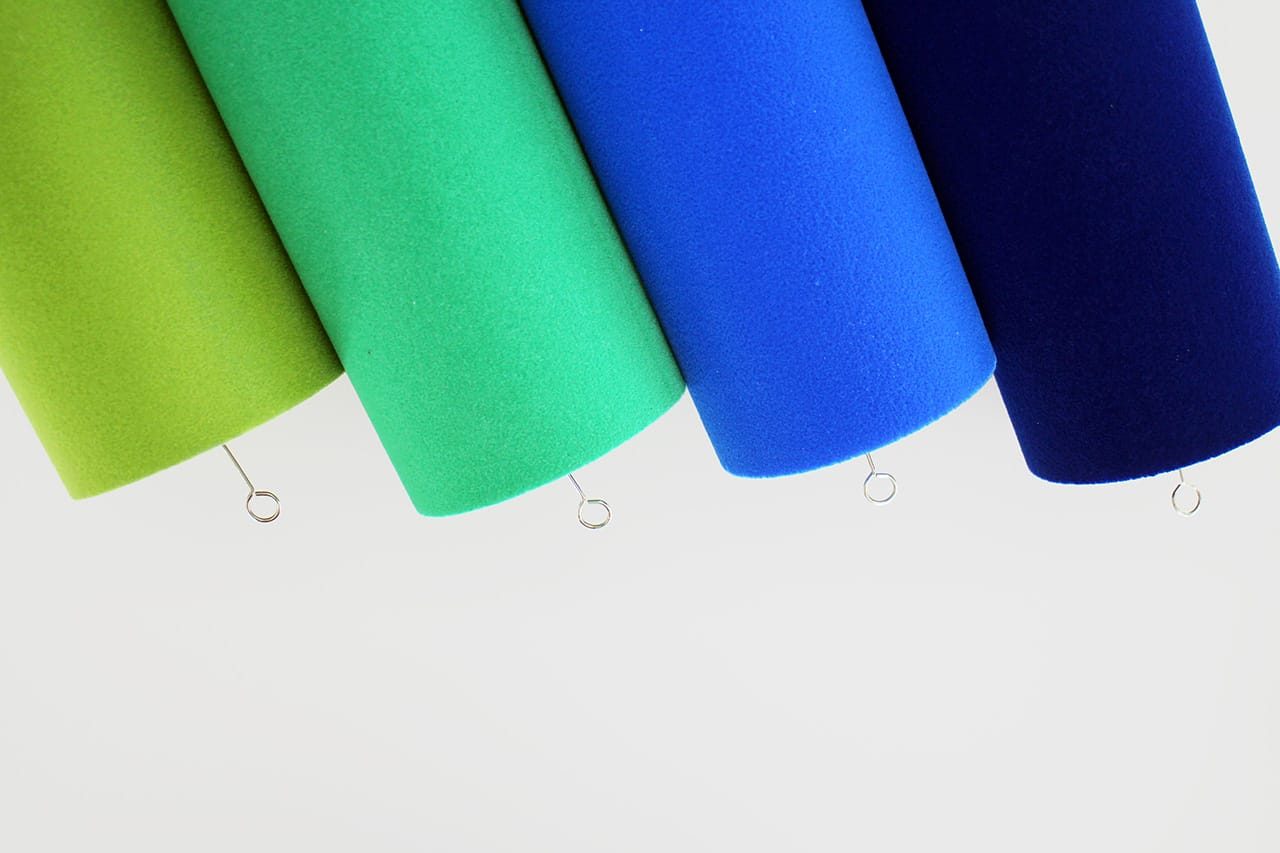 Four rolls of fabric in shades of green and blue hang side by side, each suspended by a small metal hook against a light background.