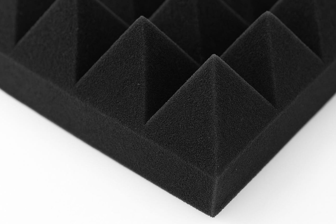 A close-up view of a black acoustic foam panel with a grid of pyramid-shaped spikes, designed to absorb sound, against a white background.