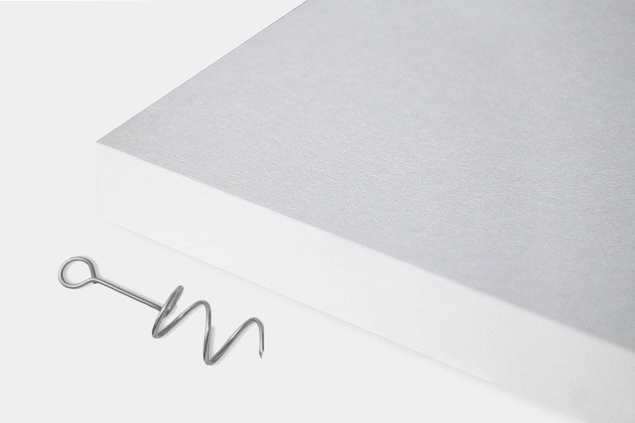 A silver corkscrew lies next to the lower right corner of a thick, white rectangular sheet or board on a plain white background.