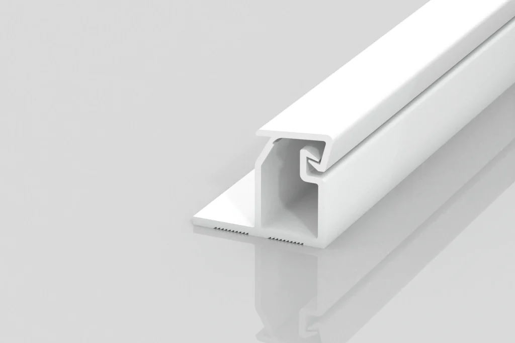 A white plastic or metal trim piece with a grooved, angular profile, lying on a light grey reflective surface. The design features an overhanging lip and an open channel along its length.