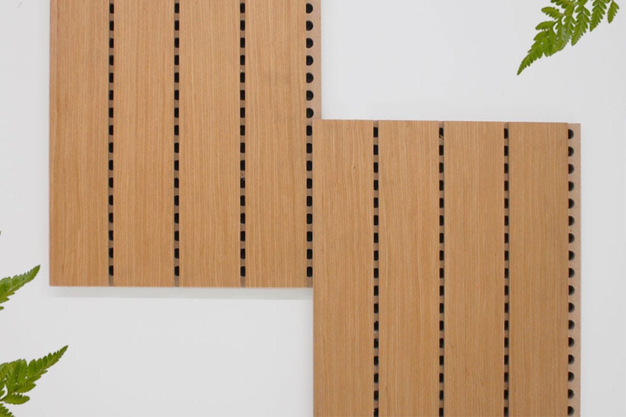 Two light wood panels with evenly spaced vertical slits are arranged on a white surface, slightly overlapping at the corner. Green fern leaves partially frame the corners of the image.