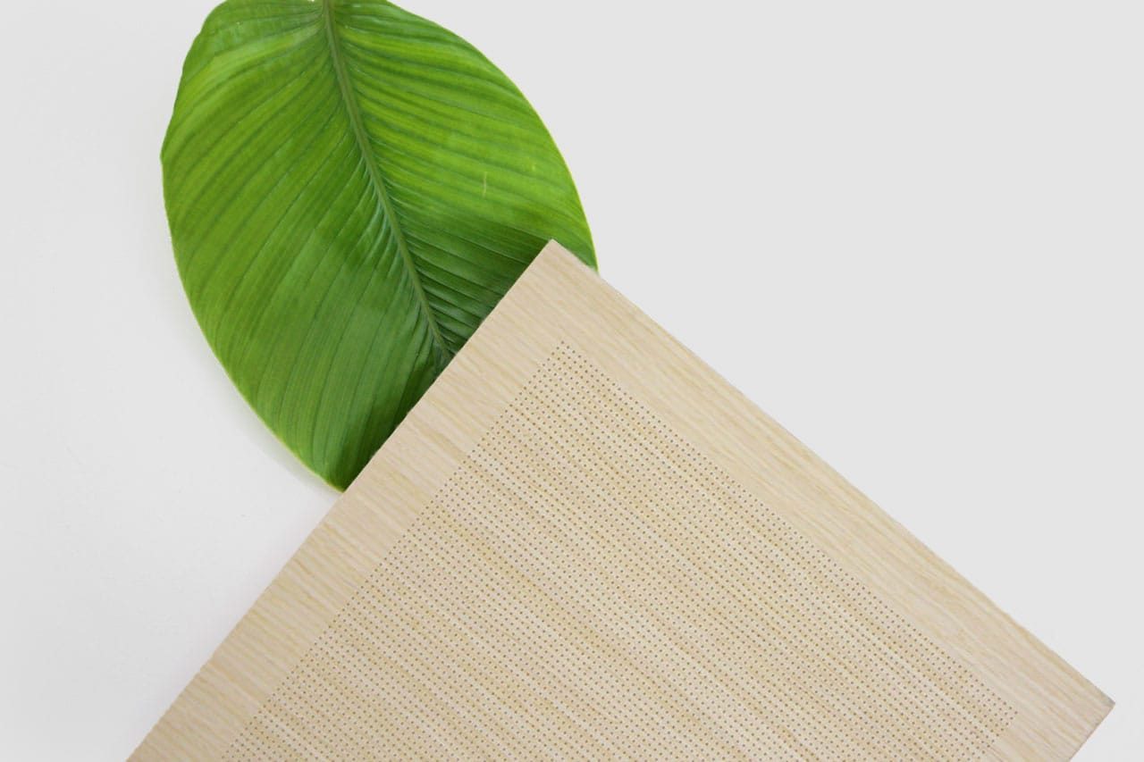 A large green leaf is placed underneath the corner of a beige woven mat on a white background. The mat covers part of the leaf, and both objects are arranged diagonally.