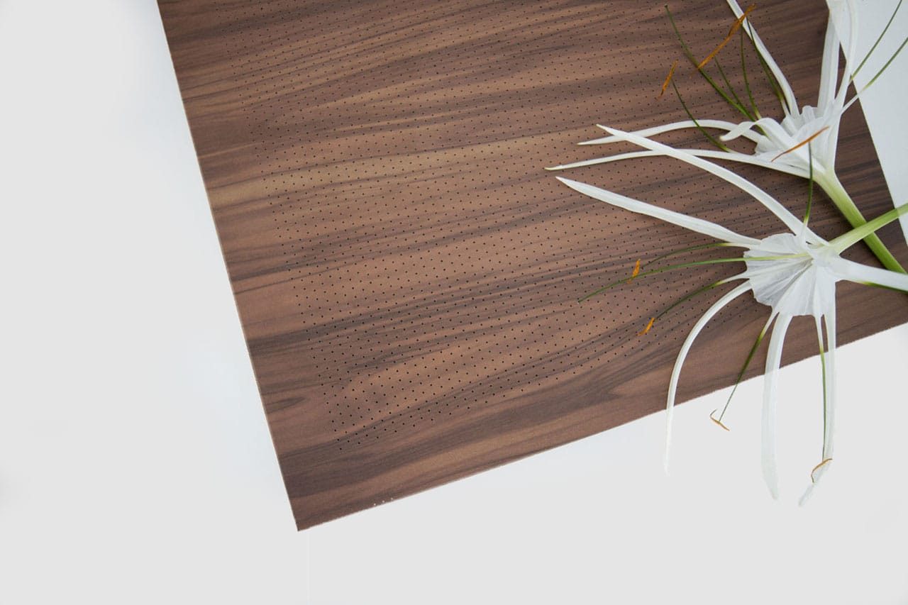 A sheet of wood veneer with small, evenly spaced perforations lies on a white surface next to delicate white spider lilies with long, thin petals and green stems.