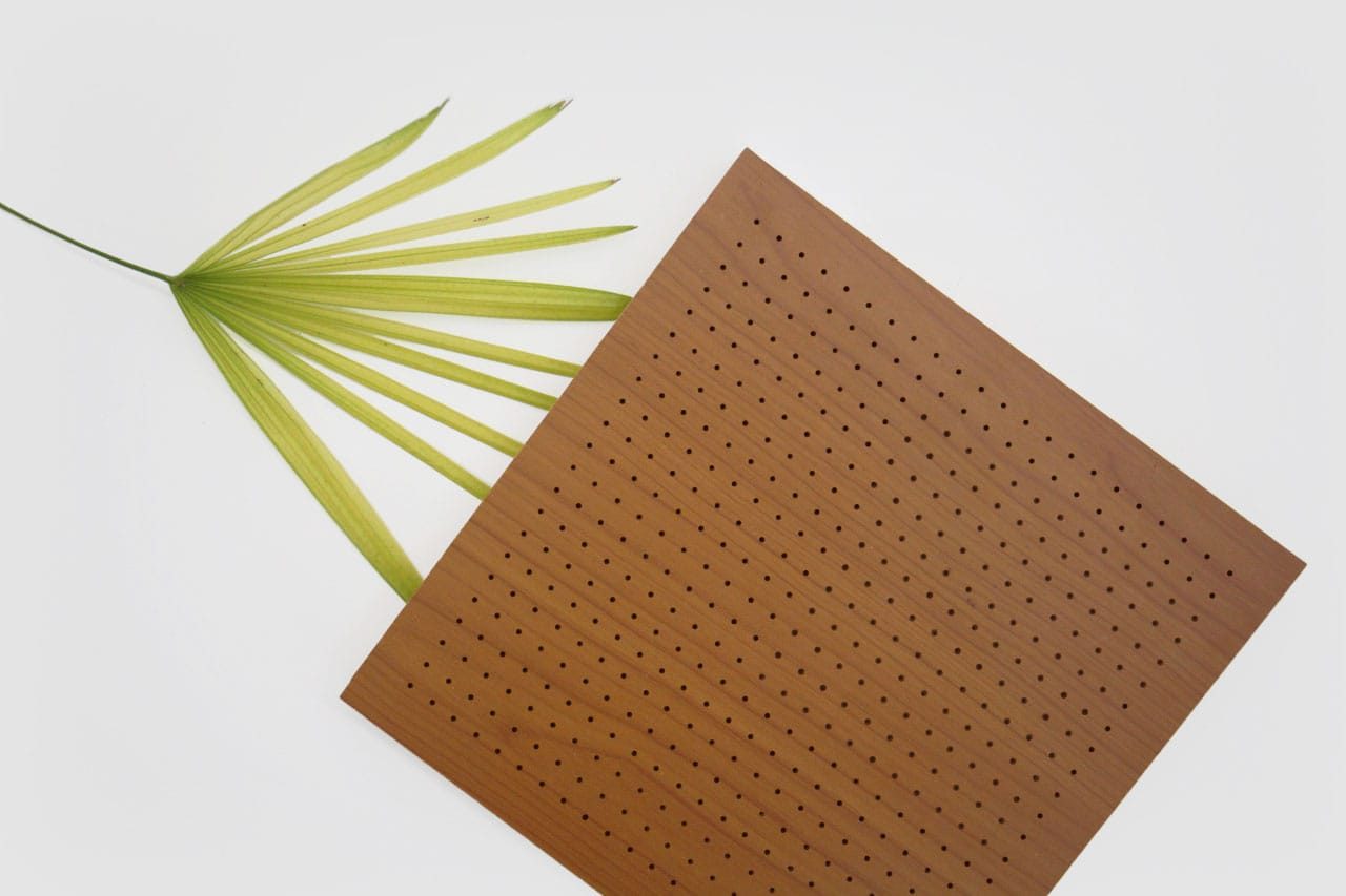 A green palm leaf and a square wooden board with evenly spaced small holes are arranged on a white background. The leaf peeks out from behind the board at an angle.
