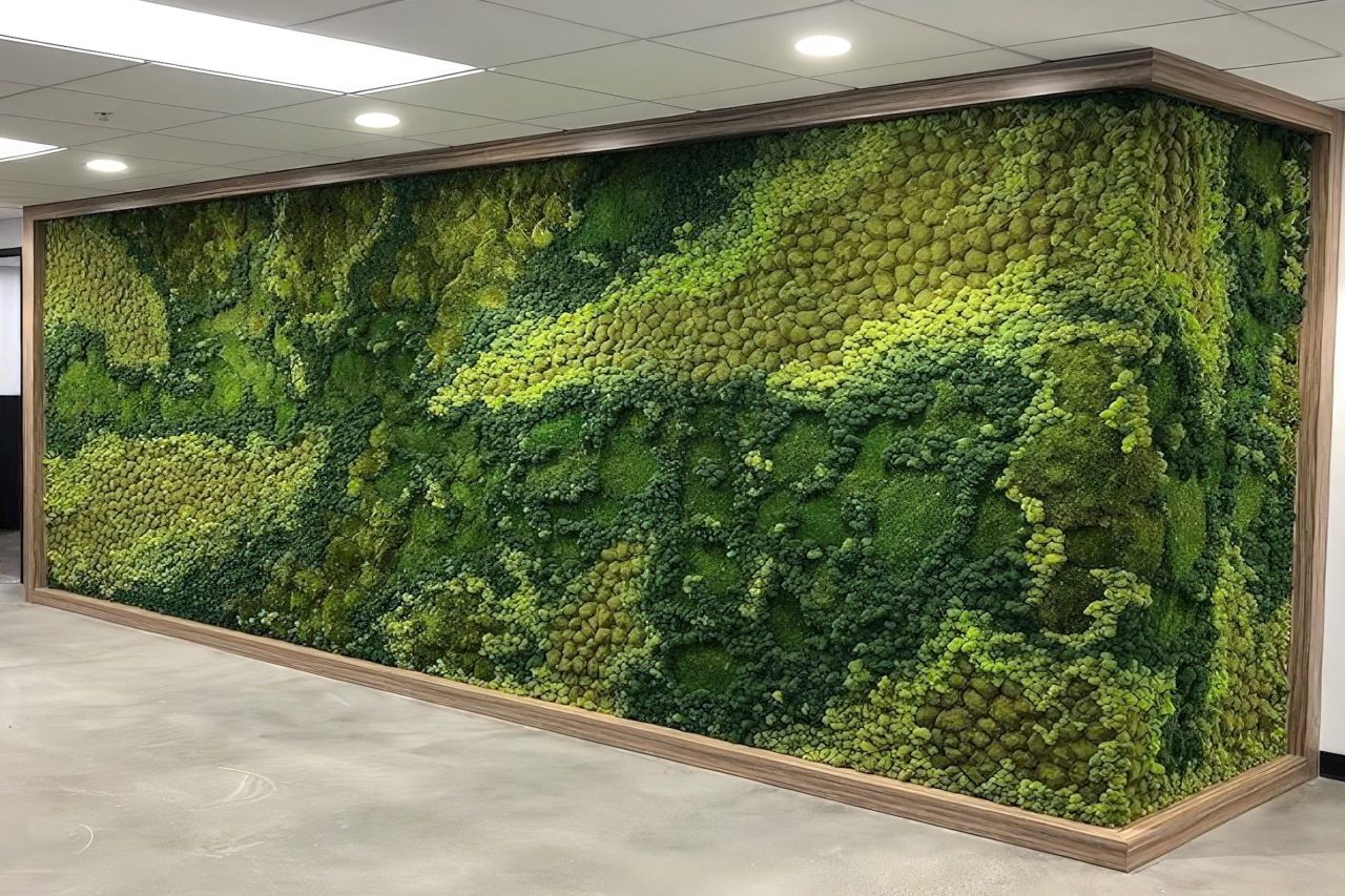 A large indoor wall is covered with lush, textured green moss in various shades and patterns, framed by wood trim. The surrounding area features a smooth, gray floor and white ceiling with recessed lighting.