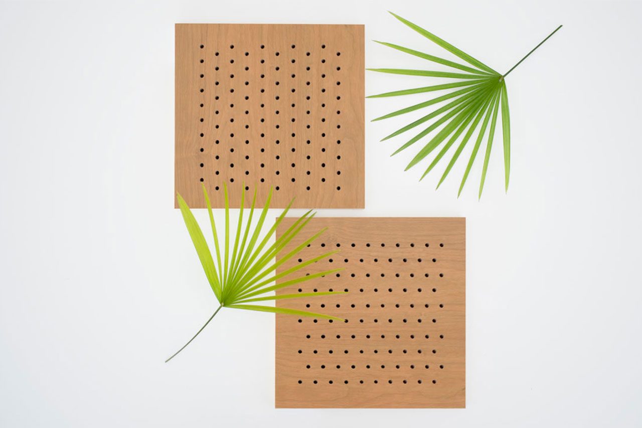 Two square wooden panels with evenly spaced small holes are arranged on a white background. Two green palm leaves are placed diagonally across and beside the panels, creating a minimalist, geometric composition.