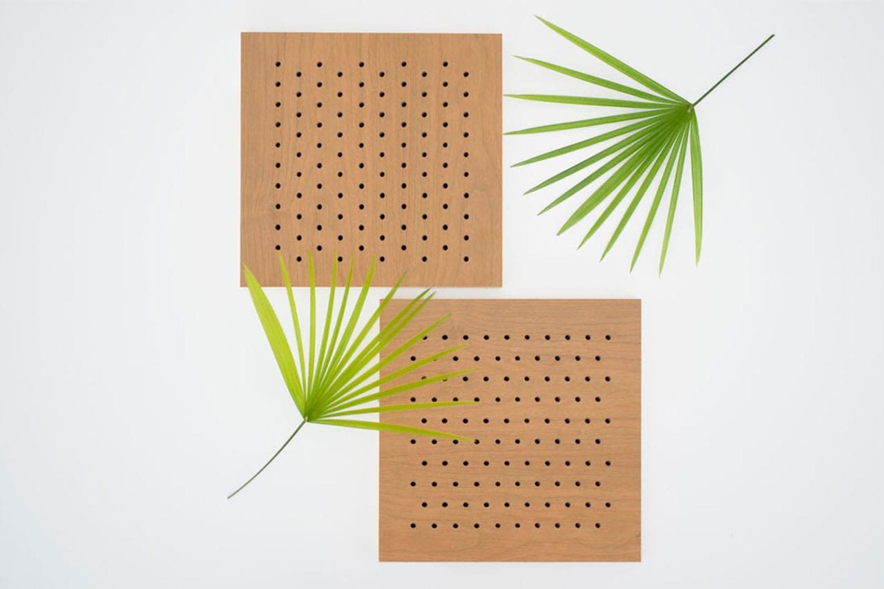 Two square wooden panels with evenly spaced small holes are arranged on a white background. Two green palm leaves are placed diagonally across and beside the panels, creating a minimalist, geometric composition.