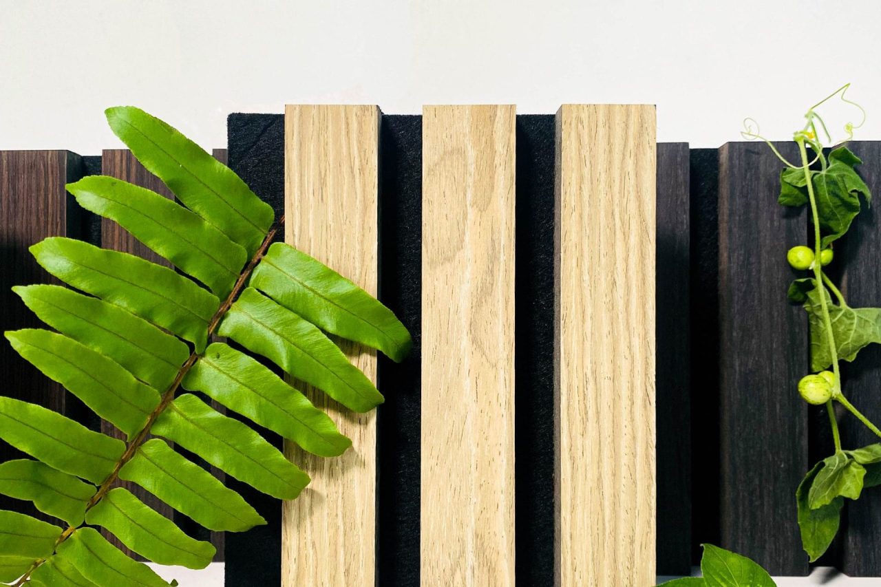 Three light wooden blocks stand vertically between dark wooden planks, with green fern leaves and small vines artfully arranged in the foreground and background against a white backdrop.