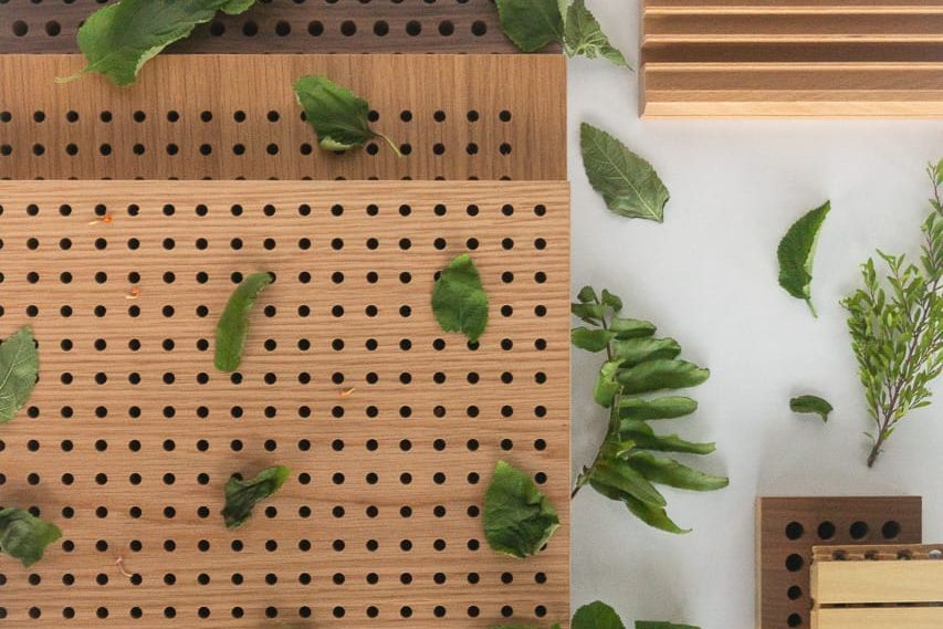 Wooden boards with holes and grooves are arranged on a white surface, surrounded by scattered green leaves and small sprigs, creating a natural and minimalistic composition.