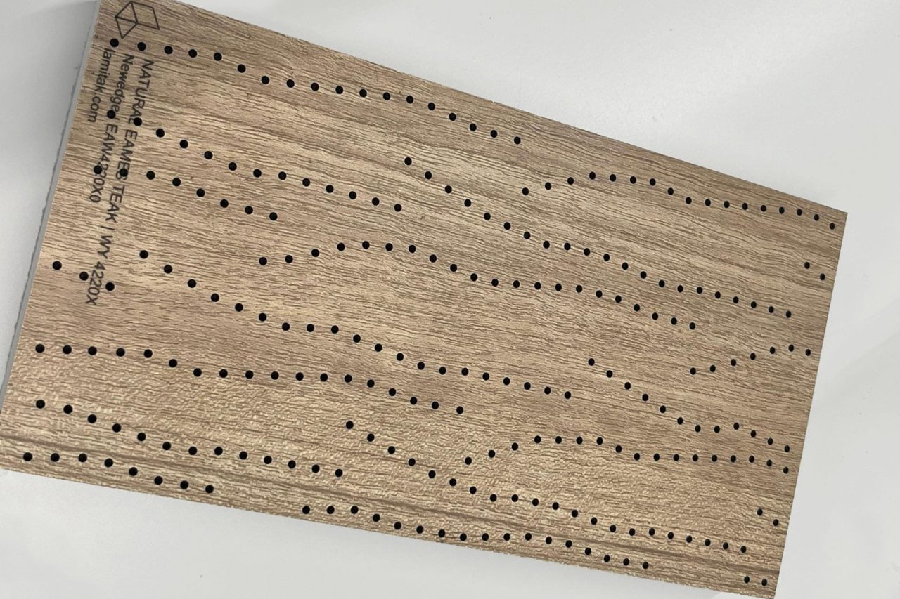 A rectangular piece of wood with evenly spaced rows of small holes forming wavy, parallel patterns. There is also engraved text in the top left corner. The background is white.
