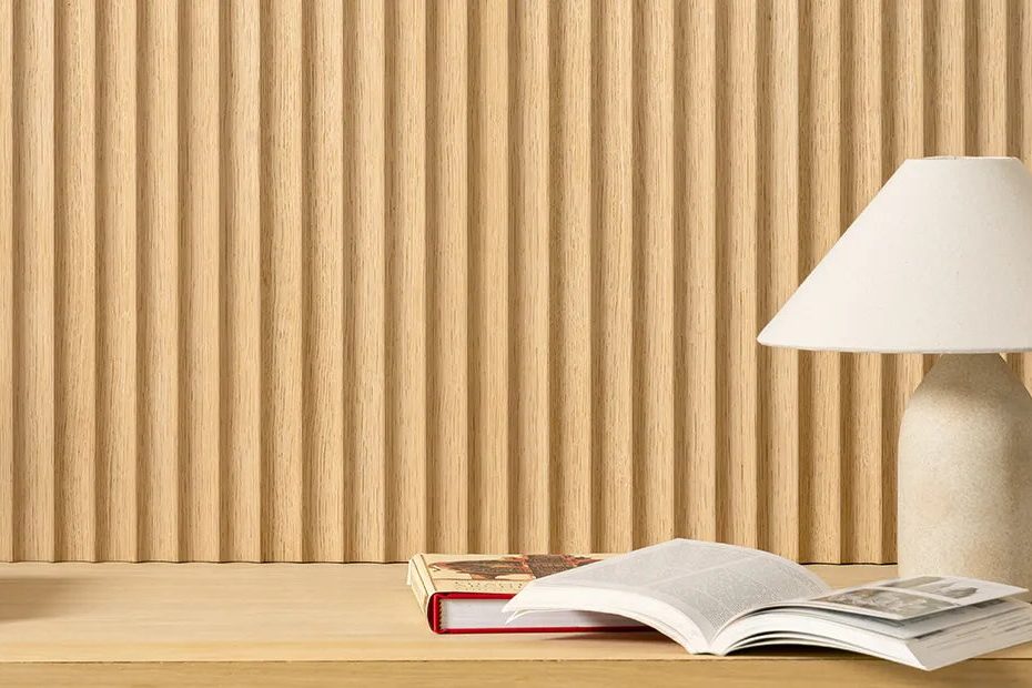 A minimalist desk with an open book, a closed book, and a beige table lamp sits in front of a textured wooden panel wall.