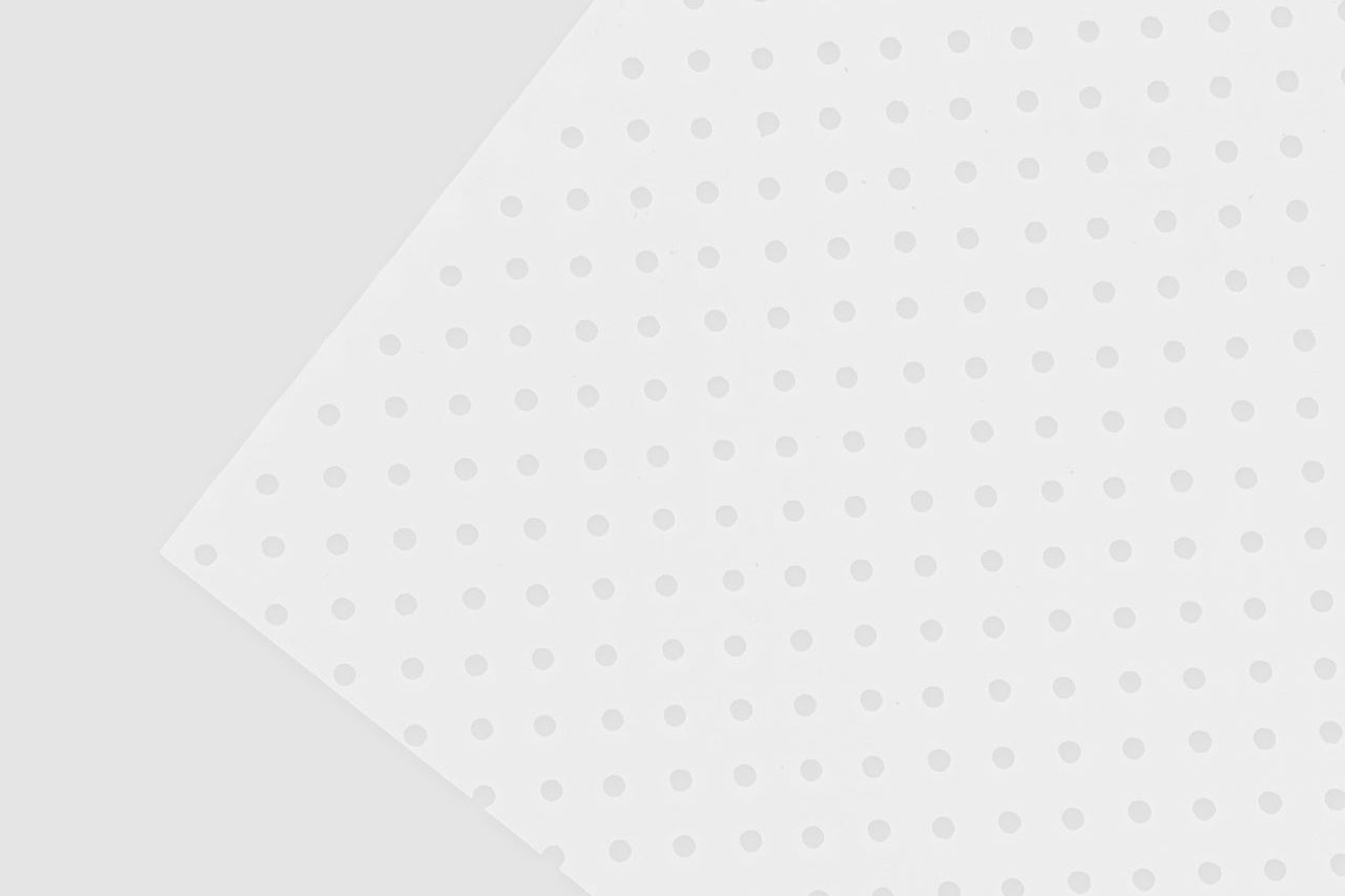 A white sheet with evenly spaced small circular holes is laid diagonally on a light gray background, creating a geometric pattern with subtle shadows and a minimalist design.